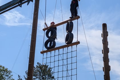 A person stands atop a high ropes course, holding a safety line, above suspended tyres and a rope net, with blue sky and trees in the background.