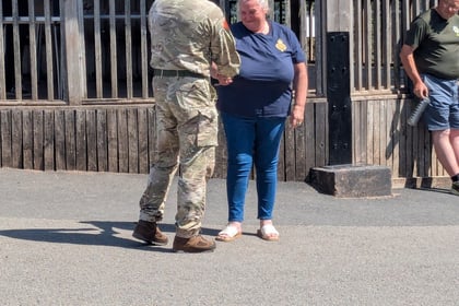 A soldier in uniform shakes hands with a woman in a blue shirt outdoors, whilst others look on from a wooden veranda in the background.