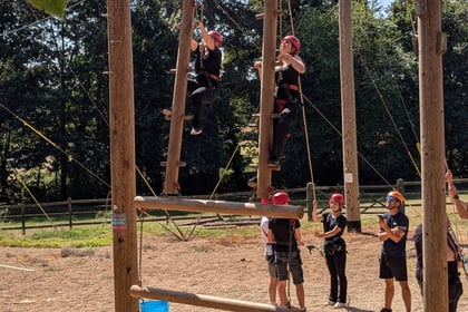 Two people climb a wooden outdoor ropes course ladder while others watch and support from below. They all wear helmets and harnesses. Trees and fencing are visible in the background.