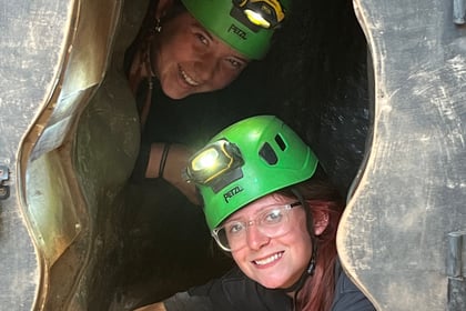 Two people wearing green helmets with head torches smile whilst posing inside a small, cave-like wooden opening. Sunlight illuminates their faces as they peer out from the enclosed space.
