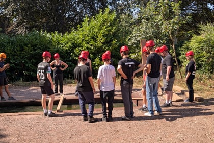 A group of people wearing red helmets stands on a dirt path near a wooden bridge over water, surrounded by green bushes and trees, during an outdoor team-building activity.