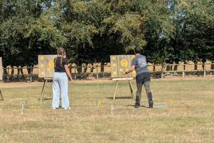 Two people stand outdoors in front of wooden boards with yellow target circles, one person throwing an object at the target whilst the other watches. Trees and a fence are in the background.