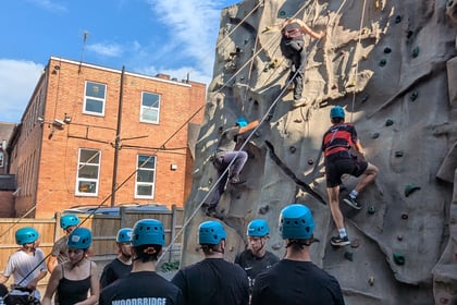 A group of people wearing blue helmets and harnesses climbs an outdoor rock wall while others watch and hold ropes, with a brick building and blue sky in the background.