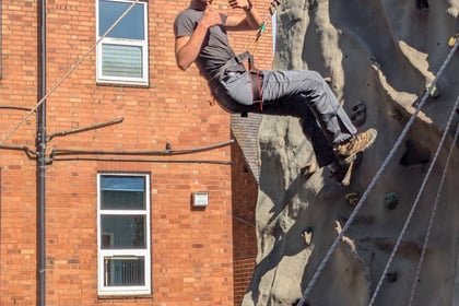 A person wearing a blue helmet and harness gives a thumbs-up whilst climbing an outdoor climbing wall. Several people in blue helmets watch from below. Brick buildings and a wooden fence are in the background.