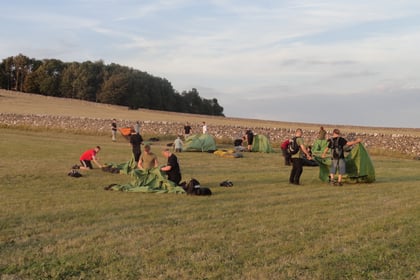 A group of people set up green tents on a grassy field with a stone wall and trees in the background, under a partly cloudy sky.
