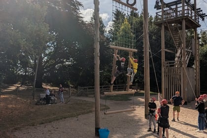 People climb a tall outdoor wooden rope course with ladders and platforms. Others watch and wait below, some wearing helmets. Trees and blue sky are in the background.
