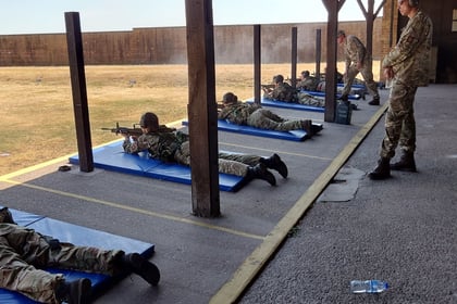 Soldiers in camouflage uniforms practise shooting while lying on blue mats at an outdoor shooting range. An instructor stands nearby overseeing the training.