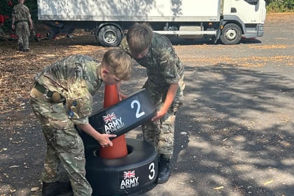 Two children in camouflage uniforms stack Army-branded weighted tyre rings marked 2 and 3 in a car park. A white lorry is parked in the background with trees and people visible.