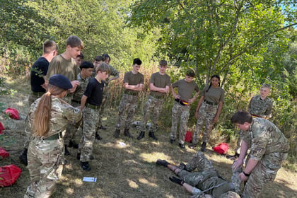 A group of young people in camouflage uniforms stand in a circle outdoors, watching as one person kneels beside another lying on the grass, likely practising a first aid or emergency drill. Trees and sunlight are visible.