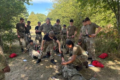 A group of young people in military uniforms practise first aid and teamwork outdoors in a grassy, wooded area on a sunny day. Some are helping others lying on the ground.