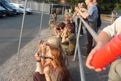 A group of people sit and stand behind a metal railing outdoors, looking and pointing at something off-camera. Some hold cameras or phones. Trees and parked cars are visible in the background.