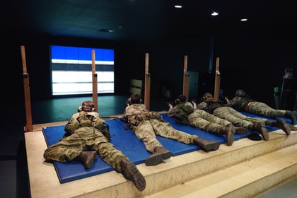 Four people in military uniforms lie prone on mats, aiming rifles at a digital shooting range screen indoors, simulating target practice.