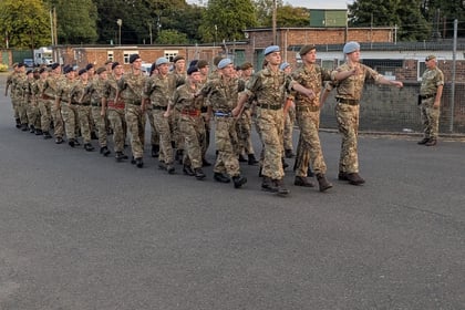 A group of uniformed soldiers marches in formation along a road, with trees and buildings in the background. An officer walks beside them, observing the group.