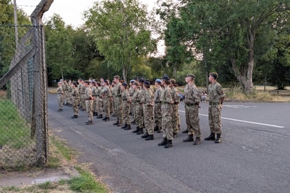 A group of people in camouflage uniforms stand in formation on a road near a chain-link fence and trees, facing a person giving instructions.