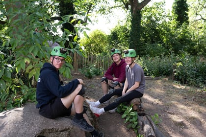 Three cadets with green helmets and knee pads on sit on a rock and a wooden log smiling at the camera