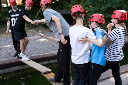 Cadets are standing either side of a water canal. There are two planks of wood across it and a cadet on the near side is reaching out to a cadet on the other to help them across.