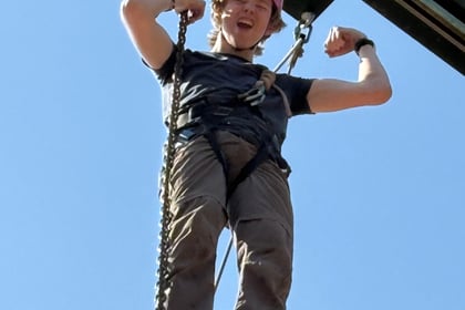 A cadet at the top of a wooden climbing structure holds on with one hand and makes a fist pump gesture with the other