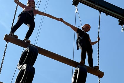 Two cadets at the top of a wooden climbing structure hold hands and look down at the camera.