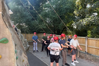 Cadets in helmets are holding ropes at the bottom of a climbing wall. You can see the legs of one cadet climbing it on the left hand side of the picture.
