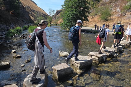 A group of cadets walking on stepping stones across a body of water. They are in civilian clothing carrying backpacks.
