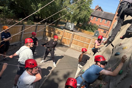 Some cadets are in a climbing wall with other cadets holding ropes to help support them. One cadet is shouting instructions to those on the wall, who are wearing blindfolds to climb.