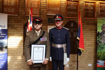 Two uniformed military officers stand in front of memorial plaques, one holding a framed certificate.