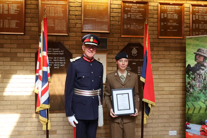 A high-ranking military officer in a formal blue dress uniform stands beside a soldier in service dress uniform who is holding a framed certificate, with military plaques and flags displayed on the brick wall behind them.