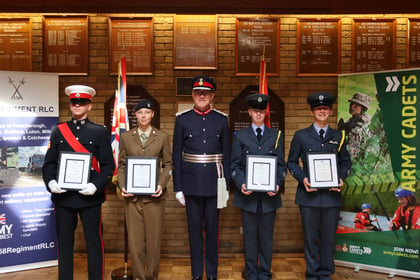 Four cadets in uniform stand in a line holding framed certificates alongside an officer in formal dress, with commemorative plaques displayed on the wall behind them.