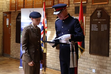 An officer in a ceremonial blue uniform presents a framed certificate to a cadet in a brown uniform.