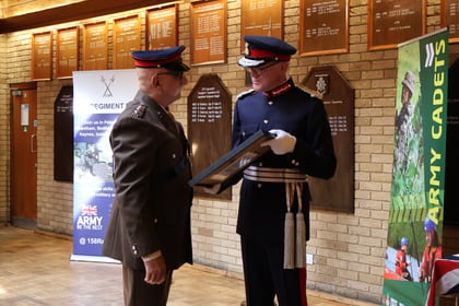 An officer in a blue military dress uniform presents a framed award to a second officer in an olive-drab uniform in front of a wall of memorial plaques.