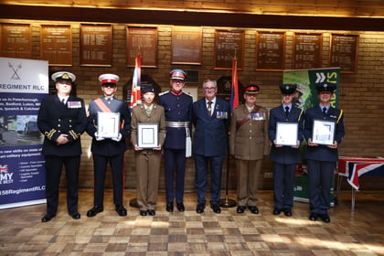 Seven military personnel in uniform, one civilian in a suit, and a guest stand in a row holding framed certificates in front of a wall of plaques.