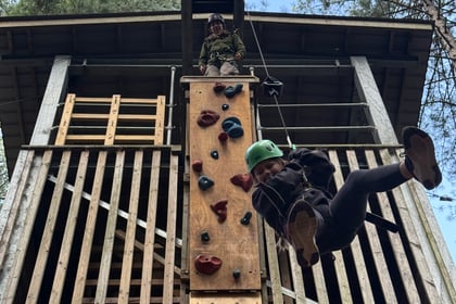 A person in a climbing helmet zips down a line attached to a wooden climbing tower, whilst an instructor watches from the top platform.