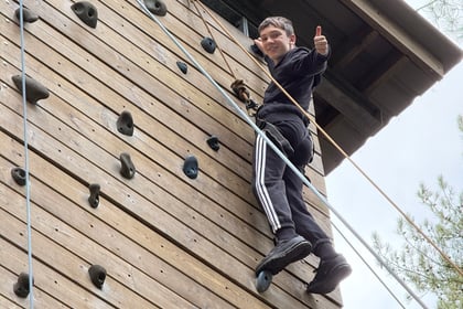A boy gives a thumbs up whilst climbing a wooden outdoor rock climbing wall, with another climber visible at the bottom left.