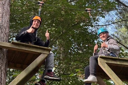 Two people wearing climbing helmets and harnesses sit on wooden platforms in the trees, each giving a thumbs-up.