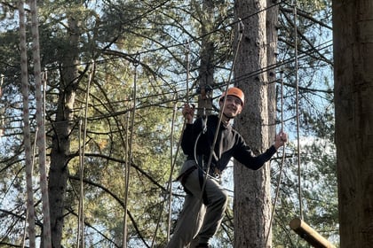 A person wearing a helmet and harness crosses a rope bridge in a forest ropes course.