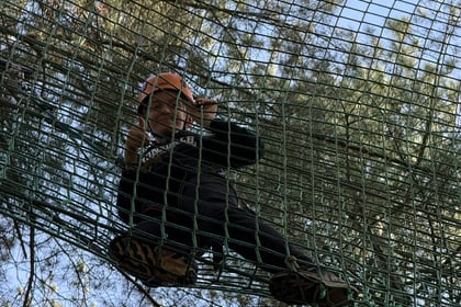A child wearing a safety helmet and harness stands in a high ropes course net suspended between trees, giving a thumbs-up.