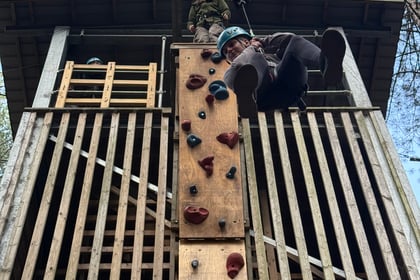 A person in a climbing harness abseils down a wooden climbing tower whilst another person supervises from the platform above.