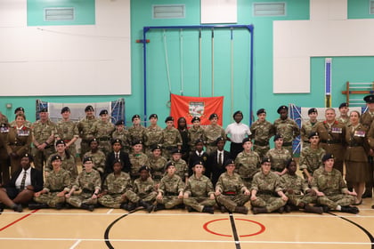 A large group of Wiltshire Army Cadet Force members and officers in uniform posing for a portrait in a gymnasium with a red company flag in the background.