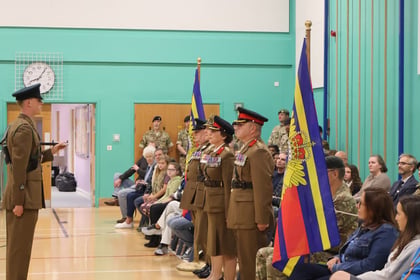 A uniformed military officer stands to attention while addressing a seated audience in a gymnasium, flanked by two ceremonial flags.