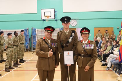 Two military officers in uniform stand on either side of a young cadet holding a certificate, with other cadets and audience members in the background of a gymnasium.