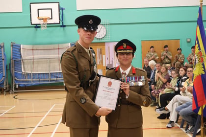 A young male cadet in uniform presents a certificate to a female officer in uniform, both standing in a sports hall with an audience in the background.