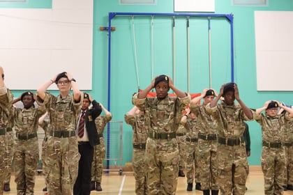 A group of youths in camouflage uniforms and dark berets stand in rows in a gymnasium, each holding their hands to the sides of their heads.