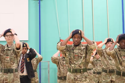 A group of young cadets in camouflage uniforms and black berets standing in rows with their hands placed on their heads.