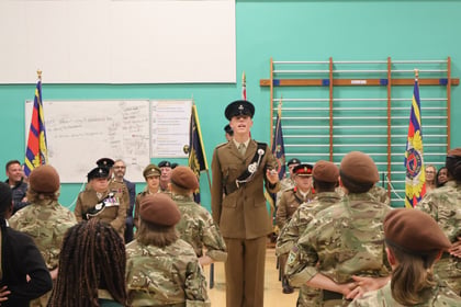 A young soldier in a brown uniform stands at the front of a room, addressing a group of cadets in camouflage uniforms and brown berets.