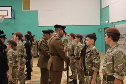 A British Army officer in uniform inspects a line of cadets in camouflage uniforms inside a gymnasium.
