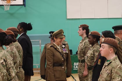 An officer in a brown uniform smiles whilst inspecting a line of Army Cadets standing in uniform in a gymnasium.