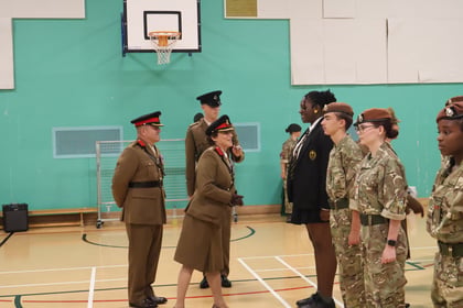 Two uniformed military officers in tan dress uniforms stand before a line of students in military cadet uniforms and one student in a school blazer in a gymnasium.