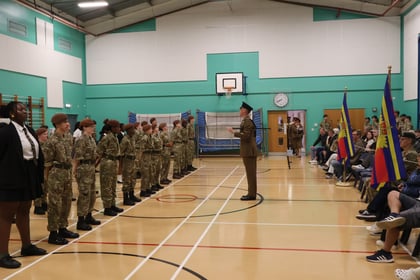 A military officer stands before a line of cadets in uniform in a gymnasium, with a seated audience watching to the right.