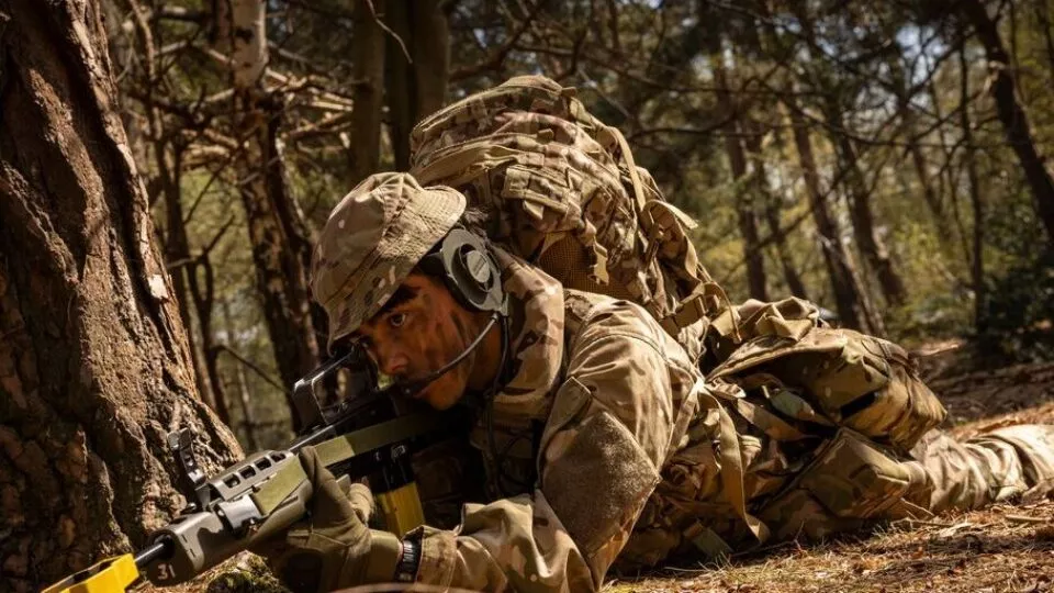 An Army cadet in camouflage uniform lies prone in a forest, aiming a rifle. The cadet wears camouflage paint, a headset, and a large rucksack, staying alert behind a tree.
