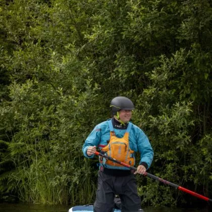 Adult volunteer on their knees on a paddle board floating across a lake wearing a blue jacket and yellow buoyancy aid and black helmet.